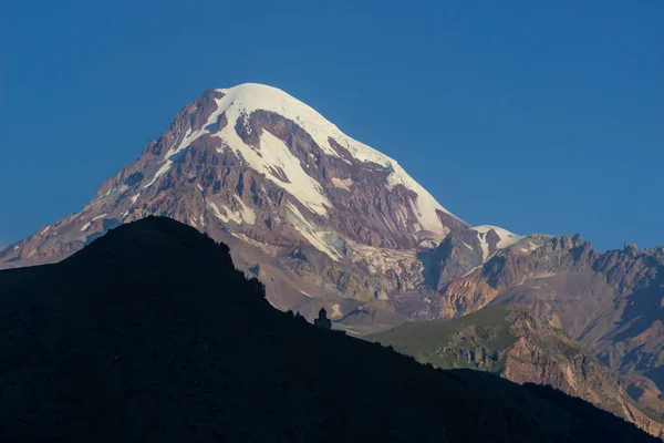 Distant view on Gergeti Trinity Church in Stepansminda, Georgia. Clear sky above the snow-capped Mount Kazbegi in the back. Sunrise, sunset Stock Image