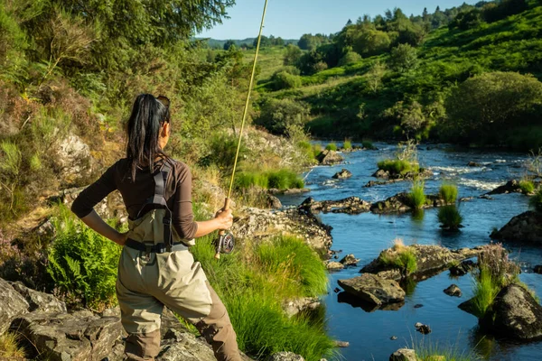 An asian female fly fisher women wearing waders and holding a rod, looking out over a rocky river in the evening sun