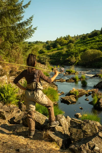 An asian female fly fisher women wearing waders and resting a rod on her shoulder, as she looks where to fish on a river in Scotland