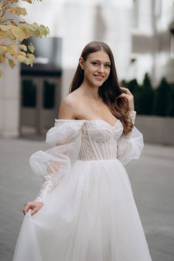 Beyaz elbiseli, uzun kıvırcık saçlı, güzel, gülümseyen gelin portresi. Uzun kollu, beyaz, omuzları, boncukları, dantelleri ve yakası olan bir gelinlik. Sevimli gelin.