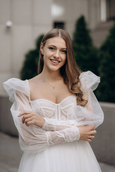 Portrait of beautiful happy bride with long curly hair in white wedding dress. Fashionable long sleeves white wedding dress with bare shoulders, beads, lace, and neckline. Adorable bride