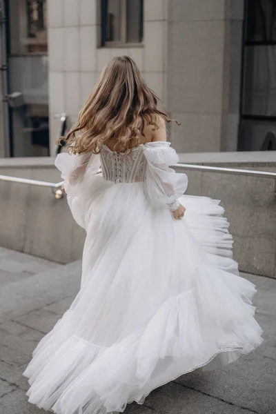 Vertical portrait of beautiful woman with long hair in white wedding dress on the city background. The bride stands with her back. Fashionable long sleeves white wedding dress with bare shoulders