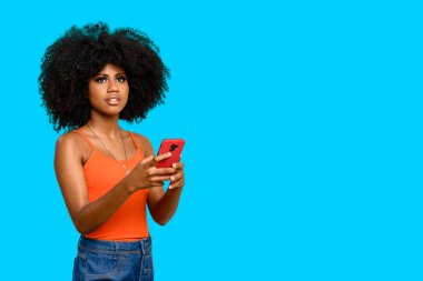 Afro saçlı genç bir kadın beyaz bir yeri işaret ediyor, bir elinde akıllı bir telefon tutuyor, mavi arka planda izole edilmiş..