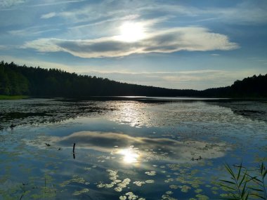 Sunset on the lake. The sun and clouds are reflected in the water. In the background a dark forest. Sunset in Masuria