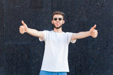 Jest, duygu, ifade ve insan konsepti - Beyaz tişörtlü, gözlüklü ve kot pantolonlu Avrupalı sakallı adam, dilini gösteriyor ve açık bir alanda siyah bir arkaplanda başparmağını kaldırıyor 