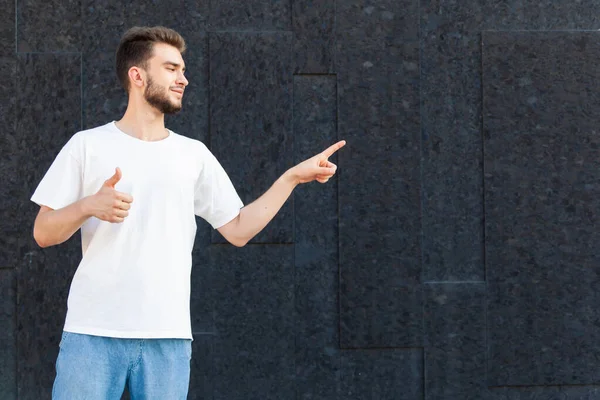 表情和人的概念 一个身穿白色T恤和牛仔裤 留着欧洲胡子的男人 他竖起大拇指 用手指在复制的空间上 用黑色背景在街上看 — 图库照片