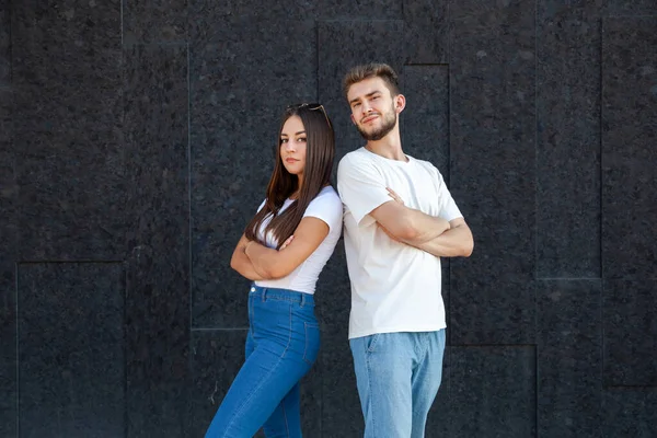 Emotions, expression and people concept - Brutal Caucasian man and woman in white T-shirts and blue jeans standing with their backs to each other and arms crossed on a black background with copy space