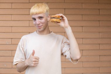 Fast food, eğlence ve insan konsepti - bej tişört giymiş Avrupalı bir adam elinde hamburgerle yüzüne yakın duruyor, başparmağını kaldırıyor ve arka plandaki duvara bakıyor.