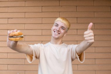Fast food, jest, eğlence ve insan konsepti bej tişört giymiş gülümseyen bir Avrupalı adam elinde hamburgerle tuğla bir arka plandaki kameraya bakıyor.