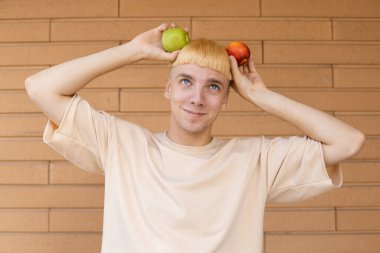 Meyveler, sağlıklı yiyecekler, organik yiyecekler ve insan konsepti - Kafkasyalı mutlu sarışın adam yeşil ve kırmızı elmalardan kulaklar yapıyor, yukarı bakıyor ve tuğla duvar arkasında bej bir tişört giyiyor