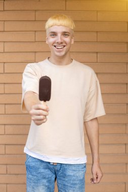 Yaz, abur cubur, şeker ve insan konsepti - elinde dondurma çubuğu tutan, kameraya gösteren ve tuğla duvar arkasında bej tişört ve kot pantolon giyen mutlu bir beyaz adam