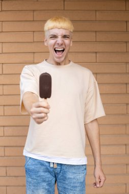 Yaz, abur cubur, şeker ve insan konsepti. Elinde dondurma çubuğu tutan beyaz bir adam, elinde bej bir tişört ve tuğla duvar arkasında kot pantolon giyiyor.