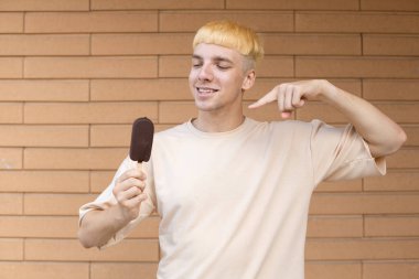 Yaz, abur cubur, şeker ve insan konsepti. Elinde dondurma çubuğu tutan, parmağıyla işaret eden, tuğla duvar arkasında bej bir tişört giyen mutlu beyaz adam.