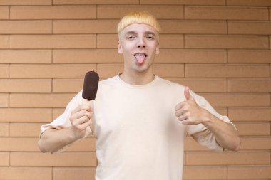 Yaz, tatlılar ve insanlar konsepti. Elinde dondurma çubuğu olan komik beyaz adam kameraya bakıyor ve duvarda bej bir tişört giyiyor.