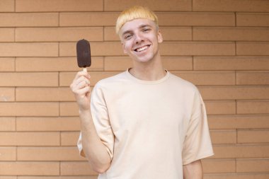 Yaz, abur cubur, şeker ve insan konsepti - yüzünde dondurma çubuğu tutan gülümseyen beyaz adam, kameraya bakıyor ve tuğla duvar arkasında bej bir tişört giyiyor