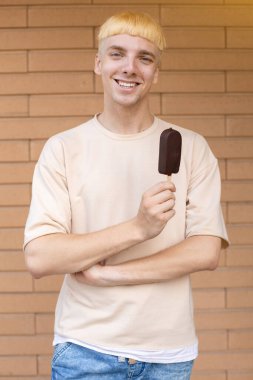 Yaz, tatlılar ve insanlar konsepti - bej tişörtlü gülümseyen beyaz bir adam ve kolları dışarıda tuğla bir duvarın arkasında çaprazlanmış bir çubukta çikolatalı dondurma tutuyor. 