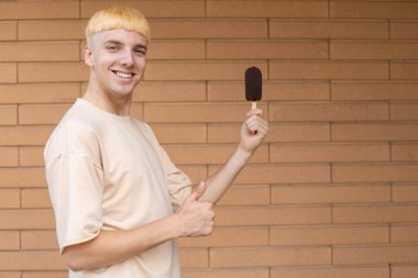 Atıştırmalık yemekler, yaz ve insan konsepti bej tişört giymiş mutlu bir beyaz adam, parmaklarını kaldırıyor ve dışarıda, tuğla duvarda, fotokopi alanı olan bir çubukta çikolatalı dondurma tutuyor.