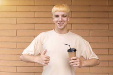 İçkiler, yaşam tarzı, tüketim, insan konsepti bej tişört giymiş gülümseyen beyaz bir adam başparmaklarını kaldırıyor ve arka planında fotokopi alanı olan tuğla bir duvarda bir fincan kahve tutuyor.