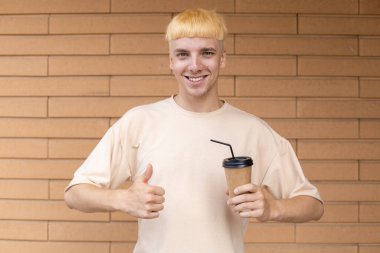 İçkiler, yaşam tarzı, jest ve insan konsepti - Bej tişört giymiş gülümseyen beyaz bir adamın yakın çekimi, başparmaklarını kaldırması ve dışarıda tuğla duvarda fotokopi alanı olan bir fincan kahve tutması