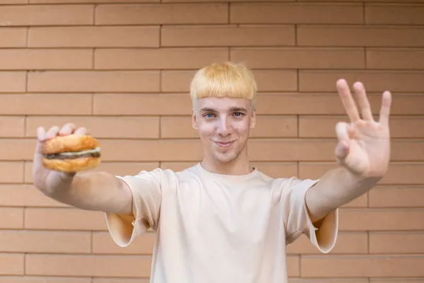休闲和人的观念 一个身穿米黄色T恤 面带微笑的欧洲男人拿着汉堡 在砖墙的背景下看着摄像机 展示出一个不错的标志 — 图库照片