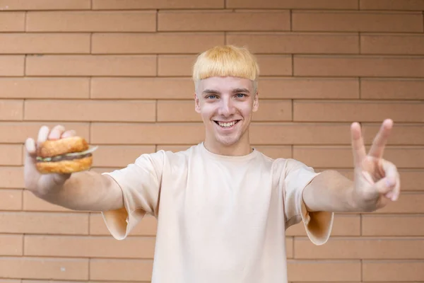 ファーストフード ジェスチャー レジャー 人々のコンセプト 彼の手にハンバーガーを保持し コピースペースとレンガの壁の背景にVの平和記号を示すベージュのTシャツを着たヨーロッパの白人男性 — ストック写真