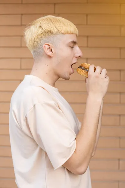 不健康的饮食和人的观念 白种人站在旁边 吃着汉堡包 穿着米黄色T恤站在砖墙的背景上 — 图库照片