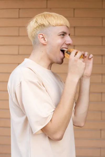 不健康的饮食和人的概念 快乐的白人金发男人侧身站立 咬着汉堡 穿着米黄色T恤在砖墙的背景上 — 图库照片