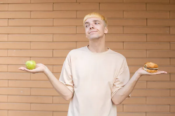 垃圾食品 健康饮食和人的概念 一个有思想的白人男人拿着一个绿色的苹果和一个汉堡包 在砖墙的背景下决定一个产品的选择 — 图库照片