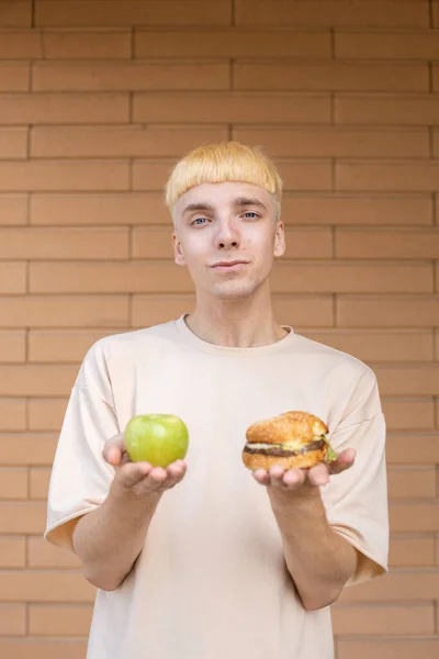 垃圾食品 健康饮食和人的概念 一个有思想的白人男人拿着一个绿色苹果和一个汉堡包 在镜头前展示他们 并在砖墙背景下提供产品选择 — 图库照片