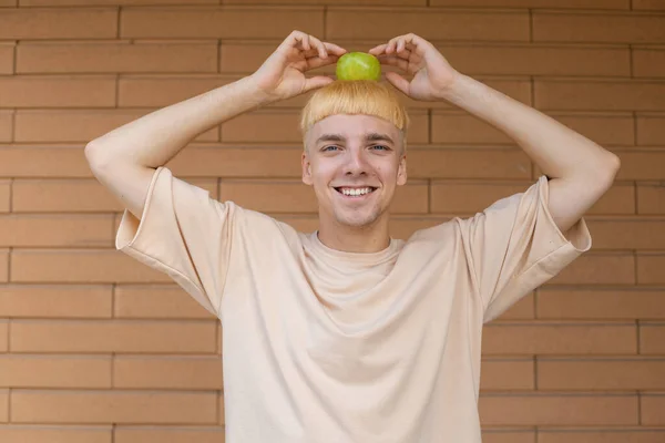 健康的な食事 人々の概念 ブロンドの白人男性がベージュのTシャツを着て レンガの壁の背景にニュートンのような頭の上に緑のリンゴでカメラを見て笑って — ストック写真