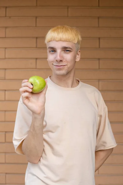 健康饮食 有机食品和人的概念 快乐的金发白种人 一只手拿着一个绿色苹果 穿着米黄色T恤 看着墙上的摄像头 — 图库照片