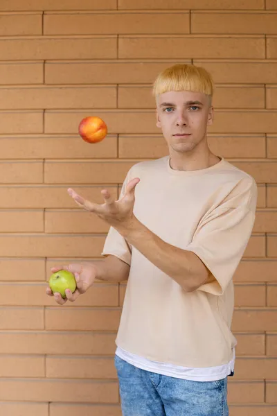 有机食品 健康饮食和人的观念 一个金发白种人在一个绿色的苹果和一个红色的油桃之间嬉戏 穿着米黄色的T恤 在砖墙的背景上看着相机 — 图库照片