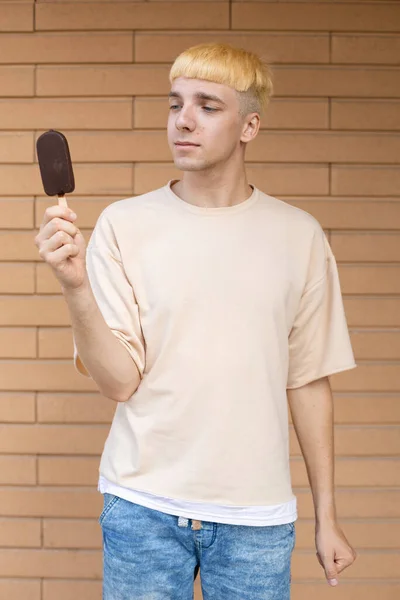 垃圾食品 糖果和人们的观念 一个快乐的白人男人手里拿着一根冰激凌棒 看着它 穿着米黄色T恤和牛仔裤在砖墙的衬托下 — 图库照片