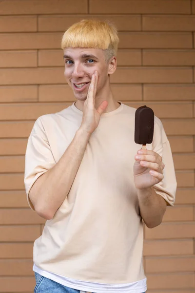 ジャンクフード お菓子 人々のコンセプト ベージュのTシャツを着たささやきの白人男性とレンガの壁の外のカメラを見て棒にチョコレートアイスクリームコーンを保持 — ストック写真