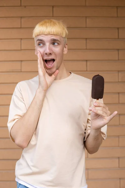ジャンクフード お菓子 人々のコンセプト ベージュのTシャツを着た悲鳴を上げる白人男性とレンガの壁に屋外の棒の上にチョコレートアイスクリームコーンを保持 — ストック写真
