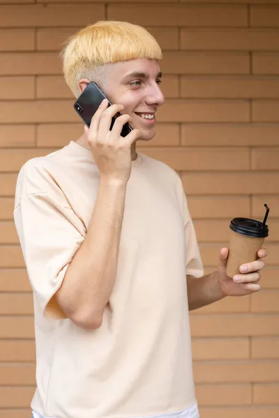 一个身穿米色T恤 面带微笑的欧洲男人 手里拿着一杯咖啡 在砖墙背景下与同事通电话 — 图库照片