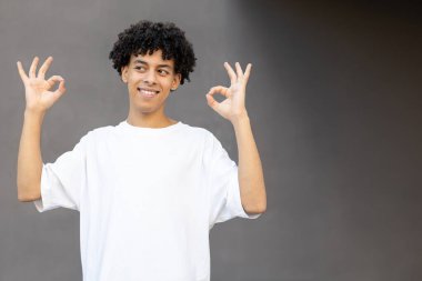 Jest, duygu, ifade ve insan konsepti - gülümseyen Amerikalı adam elleriyle iyi işaretler gösteriyor, beyaz bir tişört giyiyor ve gri bir arka planda fotokopi alanı ile dışarı bakıyor.