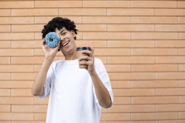 Tatlı, içki, yaşam tarzı ve insan konsepti. Elinde bir fincan kahve, elinde mavi bir çörek, kameraya bakıyor ve duvara beyaz bir tişört giyiyor.