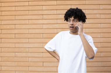 Teknoloji, yaşam tarzı ve insan konsepti. Akrabalarıyla telefonda konuşan Afro-Amerikan bir adam ve şok edici haberi tuğla bir duvarda beyaz bir tişört giyerek öğreniyor.