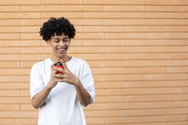 İçkiler, yaşam tarzı, tüketim ve insan konsepti. Elinde kırmızı bir fincan kahve tutan, beyaz tişört giyen ve dışarıdaki duvara bakan Afro-Amerikan adam.