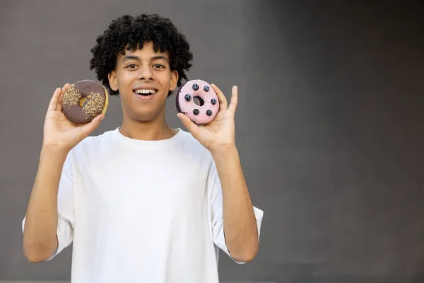娱乐和人的概念 微笑着 一个拿着巧克力和蓝莓甜甜圈的非裔美国人靠近他的脸 穿着一件灰色背景的白色T恤 带有复制空间 — 图库照片