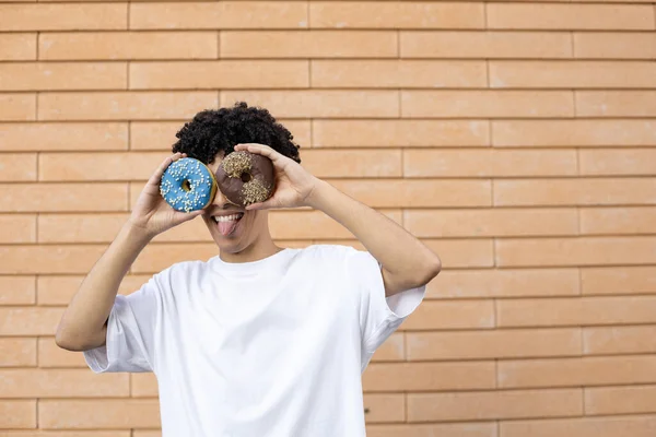 乐趣和人的概念 有趣的非裔美国人 用巧克力和蓝色甜甜圈做眼睛 展示舌头 穿着白色T恤在砖墙背景上 有复制的空间 — 图库照片