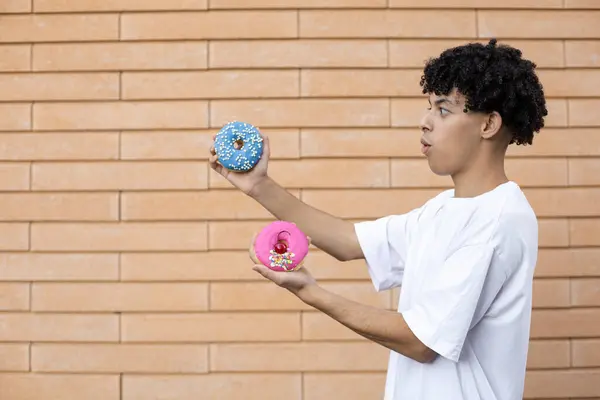 糖果和人的概念 令人惊讶的非裔美国人侧身站着 穿着一件白色T恤衫 一边拿着粉红和蓝色的甜甜圈 一边站在砖墙的背景上 — 图库照片