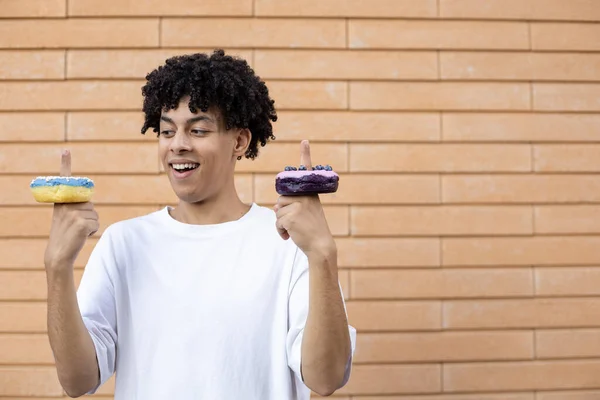 糖果和人的概念 一个快乐的非裔美国人 手指上戴着蓝色和紫色的甜甜圈 穿着白色的T恤 站在砖墙的背景上看 — 图库照片