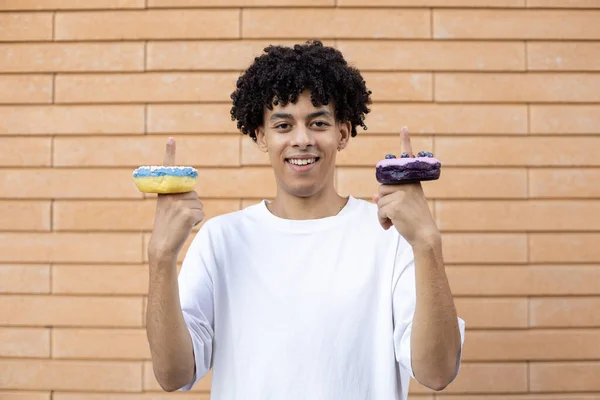 デザート お菓子 人のコンセプト 彼の指に青と紫のドーナツと幸せなアメリカ人の男は 白いTシャツを着てレンガの壁の背景にカメラを見て — ストック写真