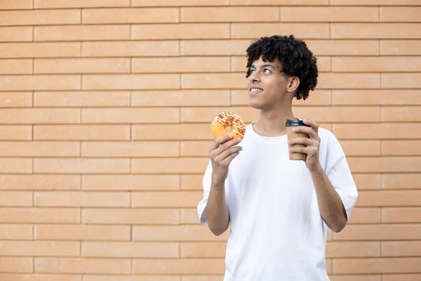 生活方式和人们的观念 满意的卷曲头发的美国男人 手里拿着橙子甜甜圈和一杯咖啡 看着复制的空间 穿着一件白色的T恤在砖墙的背景上 — 图库照片