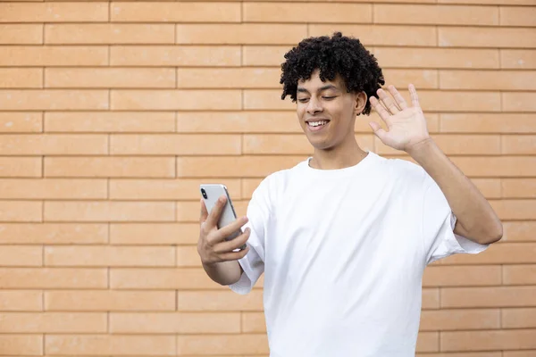 生活方式和人的观念 一个快乐的非洲裔美国人在网上学习时拿着手机 一边对着摄像机挥手致意 一边穿着一件白色的T恤衫在砖墙上 — 图库照片