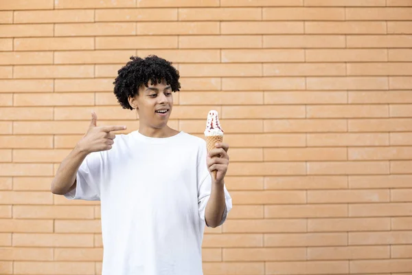 糖果和人们的观念 卷曲头发的非裔美国人 手里拿着一个冰激淋筒 用手指着它 穿着一件白色T恤放在砖墙的背景上 — 图库照片