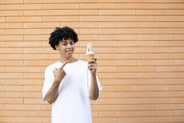 糖果和人的概念 微笑着拿着一个冰激淋筒的非裔美国人 用手指着它 看着相机 穿着一件白色的T恤在砖墙上 — 图库照片