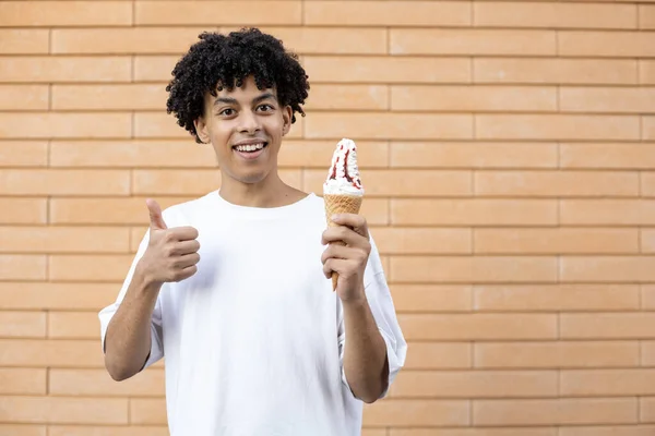 糖果和人的概念 可笑的非洲裔美国人手里拿着一个冰激淋筒 竖起大拇指 看着相机 穿着一件白色的T恤衫在砖墙上 — 图库照片